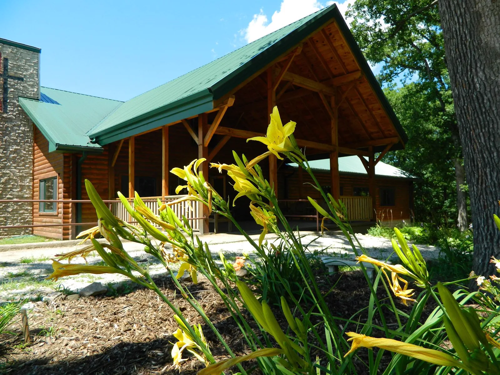 The church building, a timber lodge with a green roof and a covered porch, with daylilies flowering in front.